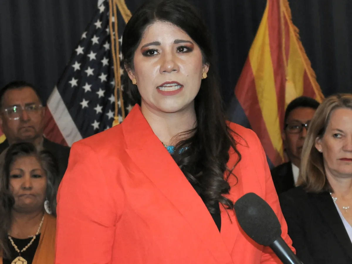 Carmen Heredia, head of Arizona Health Care Cost Containment System stands behind a microphone, making an announcement. The U.S. and Arizona flags are behind her.