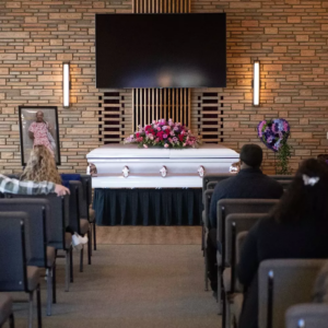 A casket sits with flowers lined on top of it. Funeral attendees sit in chairs looking at the casket.