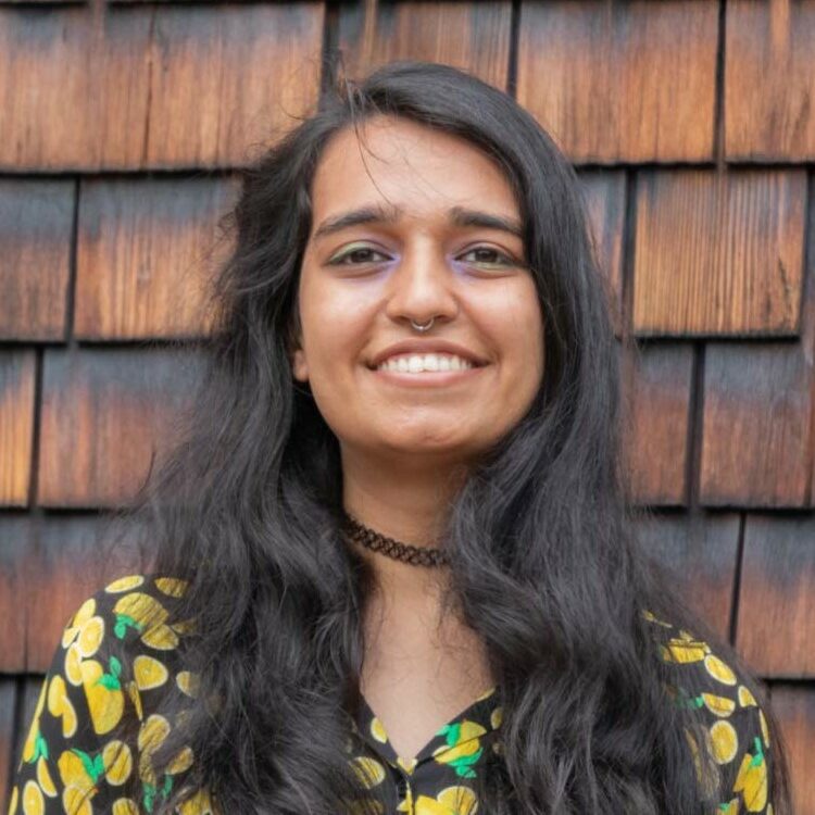 Image of Hanisha smiling with long brown hair and wearing a black and yellow shirt.