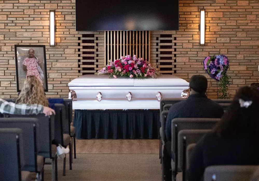 A casket sits with flowers lined on top of it. Funeral attendees sit in chairs looking at the casket.