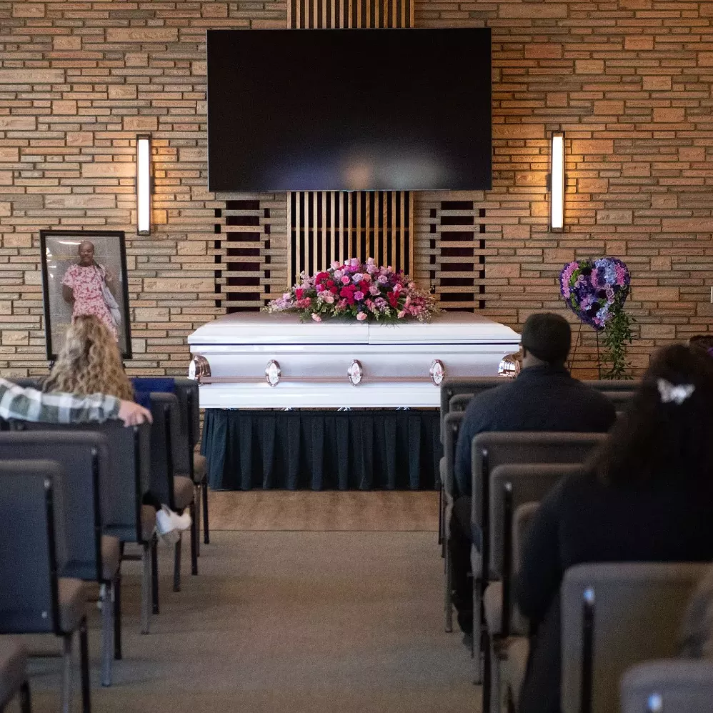 sarah_niyimbona_funeral A casket sits with flowers lined on top of it. Funeral attendees sit in chairs looking at the casket.